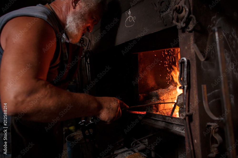 The smith artisan in his apron heats the workpiece in the burning furnace in the smith The smith artisan in his apron heats the workpiece in the burning furnace in the smith