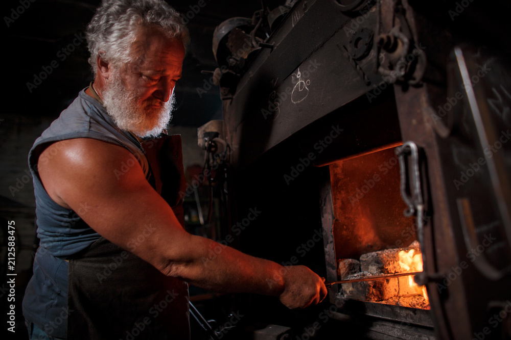 The smith artisan in his apron heats the workpiece in the burning furnace in the smith The smith artisan in his apron heats the workpiece in the burning furnace in the smith