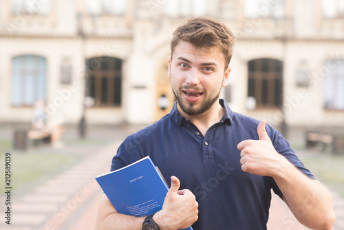 Portrait of a positive student standing with books in his hands against the background of the building and smiling. Happy man with a beard and a notebook on the background of the university. photo
