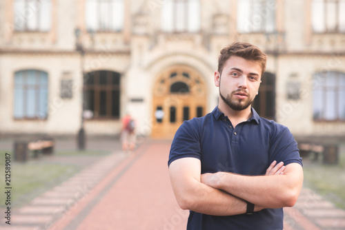 Portrait of a man with a beard and a blue T-shirt stands against the background of the building and looks at the camera. Student handsome man posing against a background of university buildings.