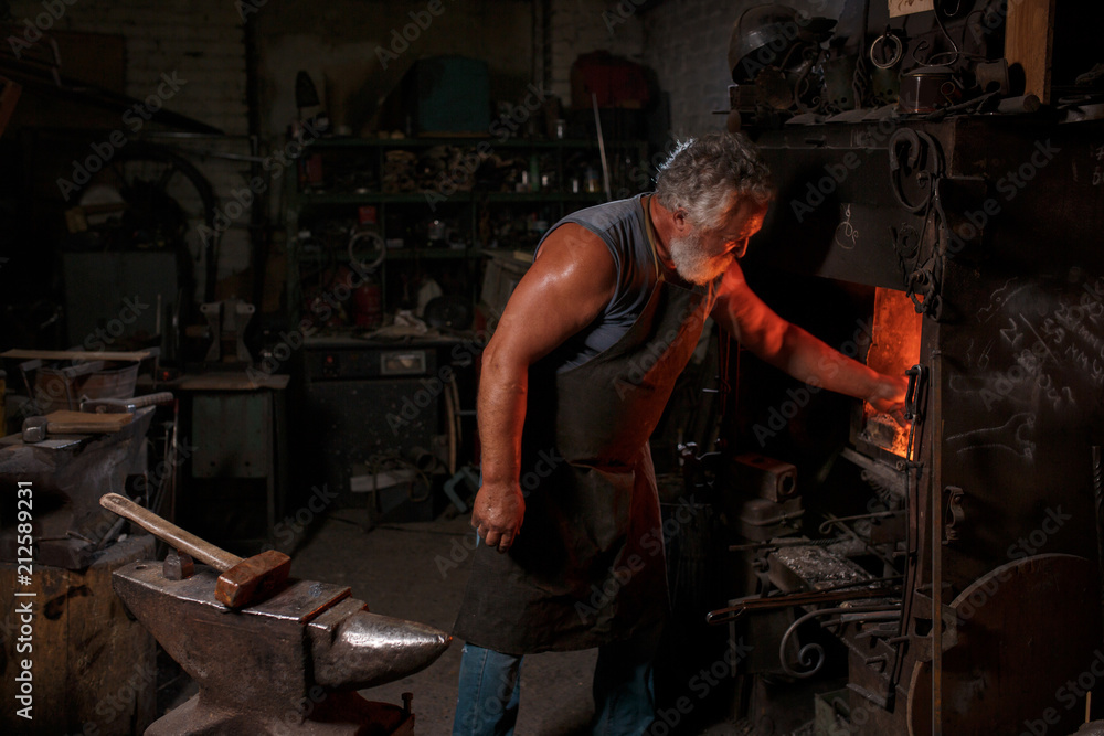 The smith artisan in his apron heats the workpiece in the burning furnace in the smith The smith artisan in his apron heats the workpiece in the burning furnace in the smith