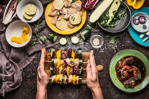 Female hands holding various homemade meat vegetables skewers for grill or bbq on rustic background with ingredients , plates and kitchen tools, top view, flat lay. Food, eating and cooking concept