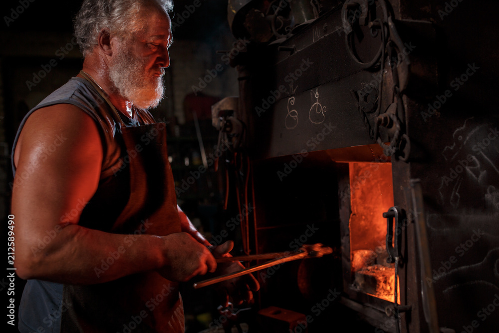 The smith artisan in his apron heats the workpiece in the burning furnace in the smith The smith artisan in his apron heats the workpiece in the burning furnace in the smith