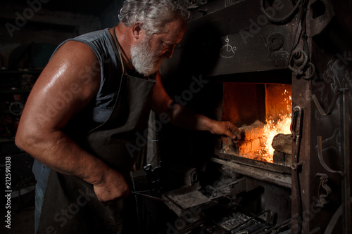 The smith artisan in his apron heats the workpiece in the burning furnace in the smith