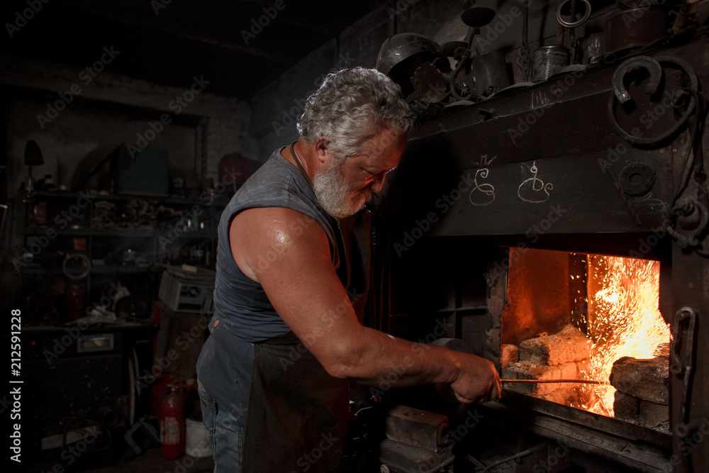 Φωτογραφία The smith artisan in his apron heats the workpiece in the burning furnace in the