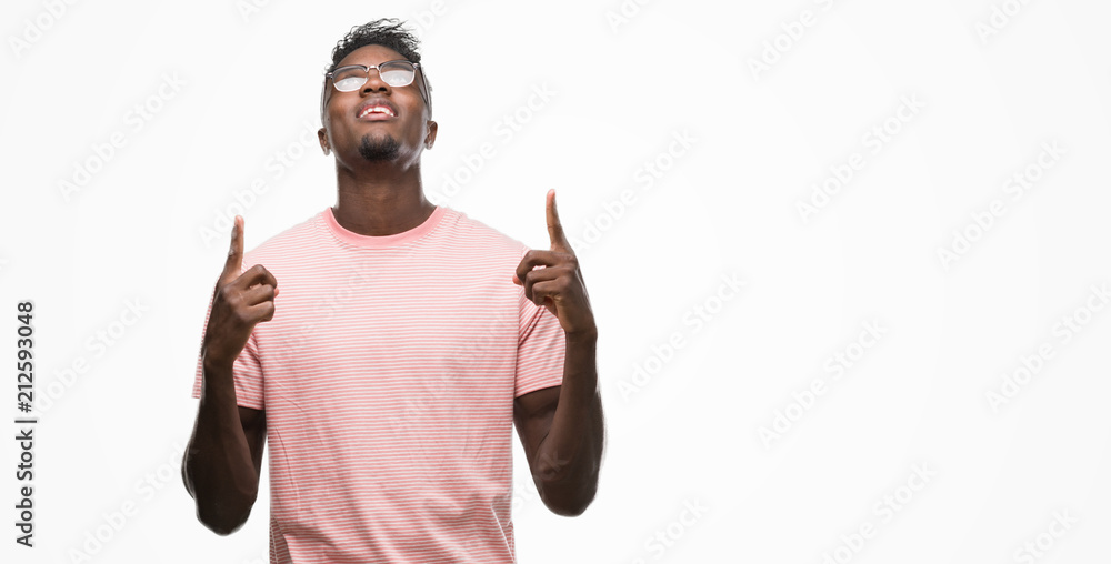 Young african american man wearing pink t-shirt amazed and surprised looking up and pointing with fingers and raised arms. Young african american man wearing pink t-shirt amazed and surprised looking up and pointing with fingers and raised arms.