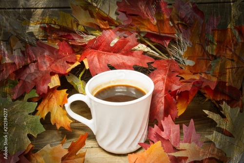 A single hot cup of coffee encircled by leaves in autumn colors on a wooden surface with a texture applied to the fringes of the image.