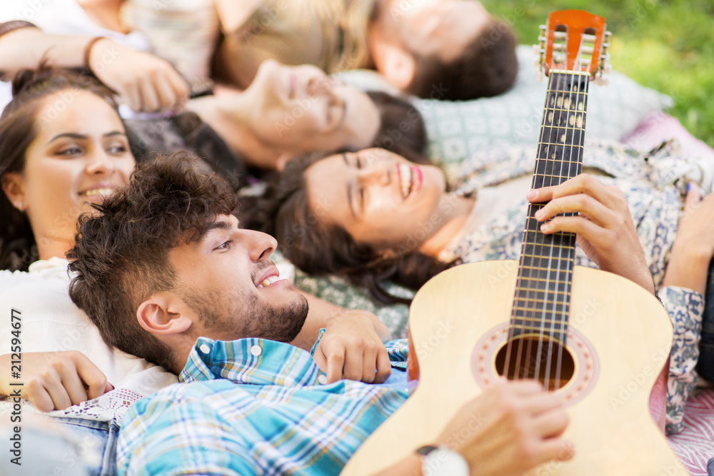 Fototapeta premium friendship, leisure and summer concept - group of happy smiling friends playing guitar and chilling on picnic blanket