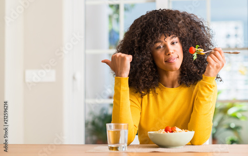 African american woman eating pasta salad at home pointing with hand and finger up with happy face smiling