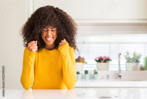African american woman wearing yellow sweater at kitchen excited for success with arms raised celebrating victory smiling. Winner concept.