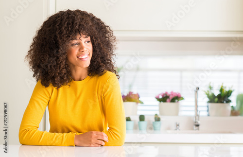 African american woman wearing yellow sweater at kitchen looking away to side with smile on face, natural expression. Laughing confident.