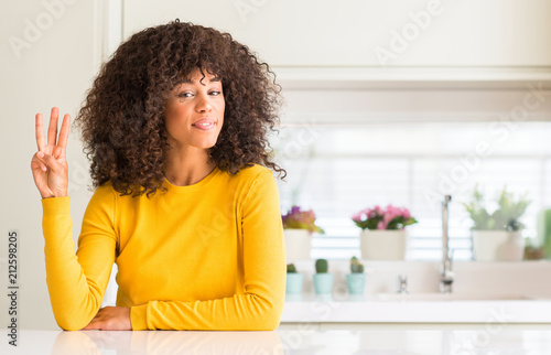 African american woman wearing yellow sweater at kitchen showing and pointing up with fingers number three while smiling confident and happy.