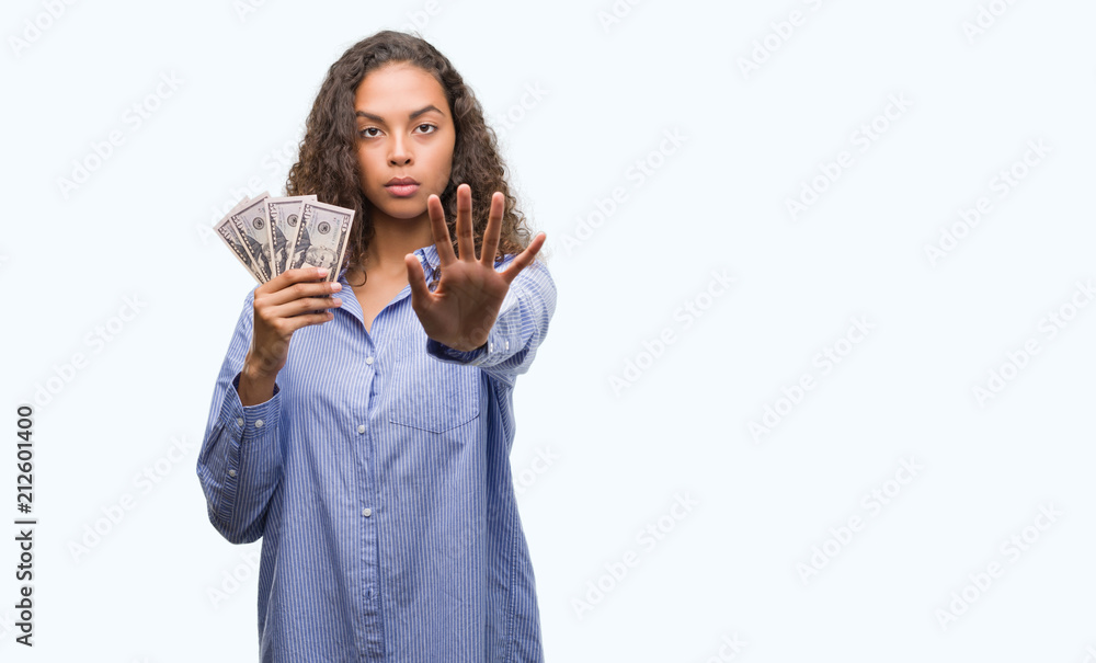 Young hispanic woman holding dollars with open hand doing stop sign with serious and confident expression, defense gesture Young hispanic woman holding dollars with open hand doing stop sign with serious and confident expression, defense gesture