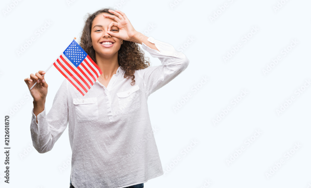 Young hispanic woman holding flag of United Estates of America with happy face smiling doing ok sign with hand on eye looking through fingers