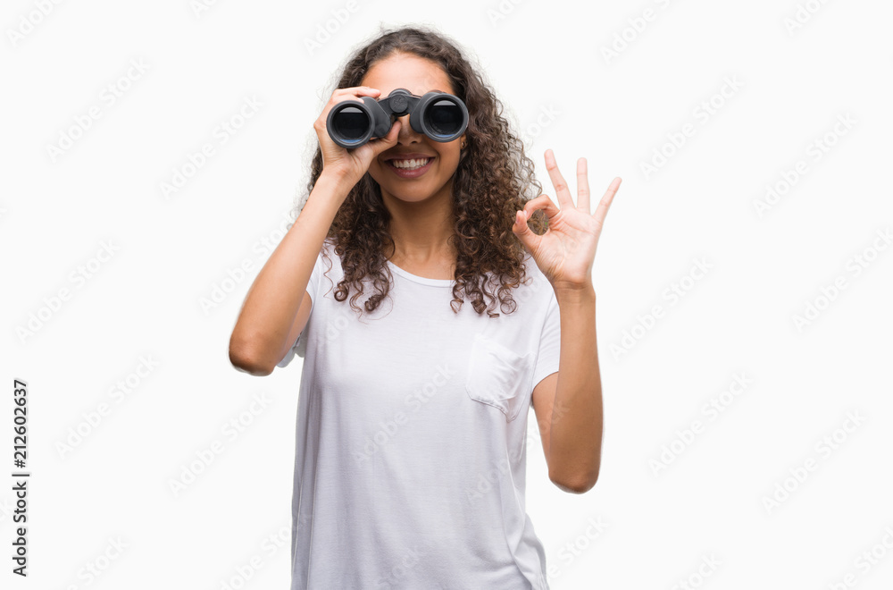 Young hispanic woman looking through binoculars doing ok sign with fingers, excellent symbol Young hispanic woman looking through binoculars doing ok sign with fingers, excellent symbol