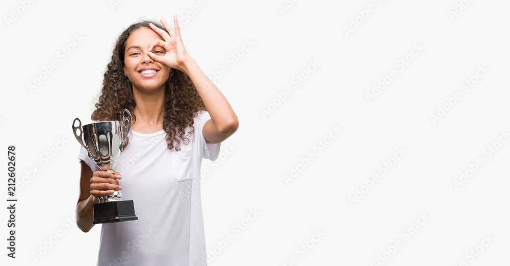 Young hispanic woman holding trophy with happy face smiling doing ok sign with hand on eye looking through fingers Young hispanic woman holding trophy with happy face smiling doing ok sign with hand on eye looking through fingers