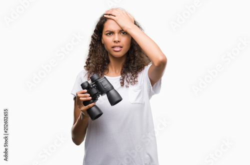 Young hispanic woman looking through binoculars stressed with hand on head, shocked with shame and surprise face, angry and frustrated. Fear and upset for mistake.