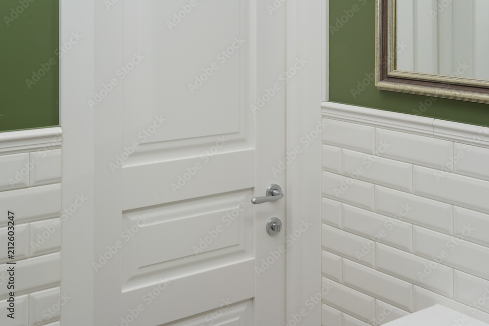 White door in the bathroom toilet room. Background green painted wall covered with decorative ceramic tiles with white glossy bricks. Interior details close-up. White door in the bathroom toilet room. Background green painted wall covered with decorative ceramic tiles with white glossy bricks. Interior details close-up.