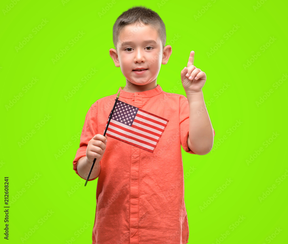 Dark haired little child holding United States flag surprised with an idea or question pointing finger with happy face, number one Dark haired little child holding United States flag surprised with an idea or question pointing finger with happy face, number one