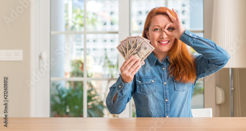 Redhead woman holding dollar bank notes at home with happy face smiling doing ok sign with hand on eye looking through fingers