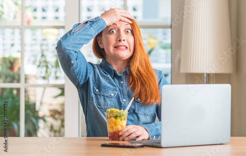 Redhead woman using computer laptop eating fruit at home stressed with hand on head, shocked with shame and surprise face, angry and frustrated. Fear and upset for mistake.