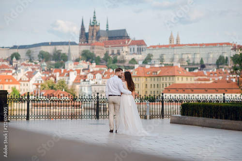 Rear view of beautiful wedding couple of groom and bride in gorgeous dress with a loop hugging and kissing in front of scenic city view of Prague, Czech Rebuplic