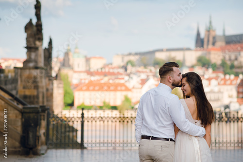 Close up back view of beautiful wedding couple of groom and bride in gorgeous dress with a loop. Man kising woman on forehead in front of scenic city view and Charles bridge in Prague, Czech Rebuplic