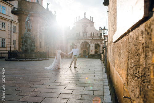 Canvas Print Romantic wedding couple of groom and bride in gorgeous dress holding hands while walking the old street of Prague