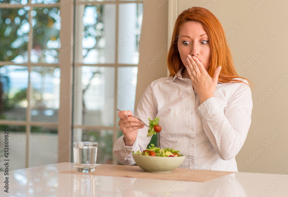 Redhead woman eating fresh green salad at home cover mouth with hand shocked with shame for mistake, expression of fear, scared in silence, secret concept