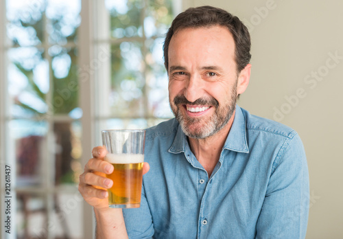 Middle age man drinking beer with a happy face standing and smiling with a confident smile showing teeth
