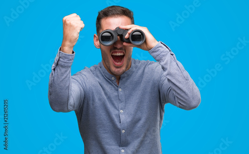 Handsome young man looking through binoculars annoyed and frustrated shouting with anger, crazy and yelling with raised hand, anger concept photo