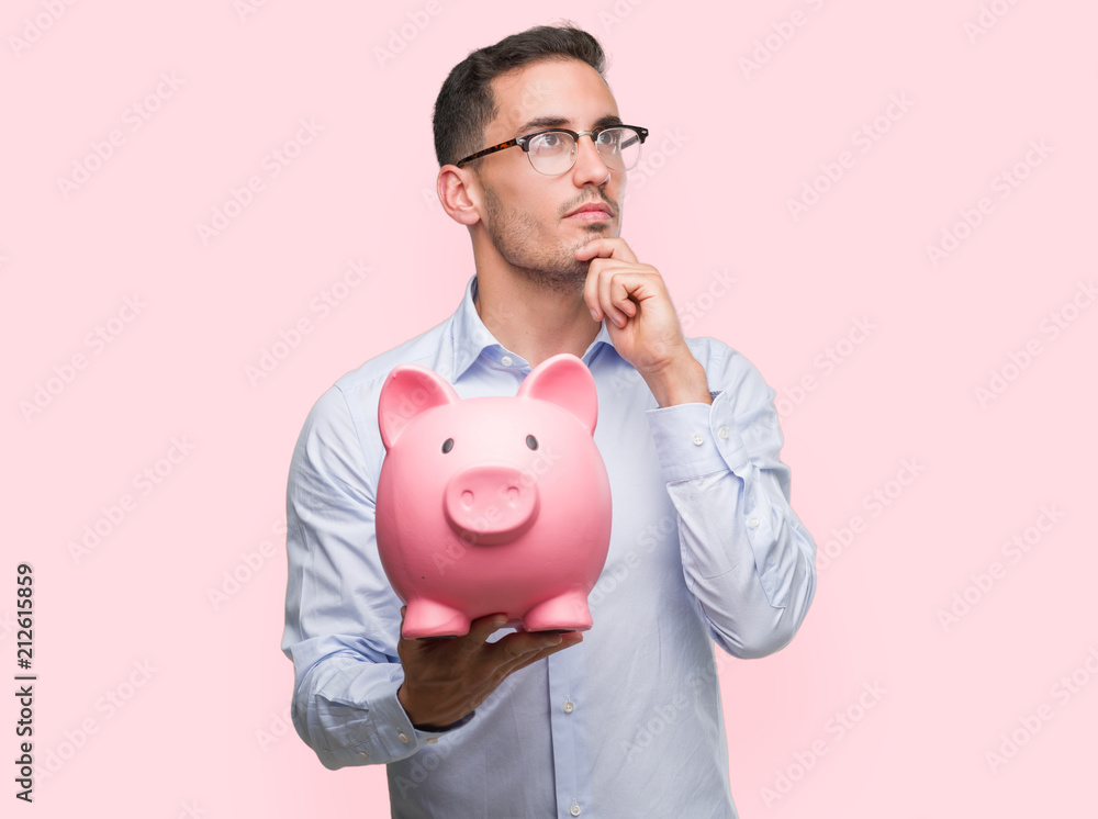 Handsome young man holding a piggy bank serious face thinking about question, very confused idea Handsome young man holding a piggy bank serious face thinking about question, very confused idea