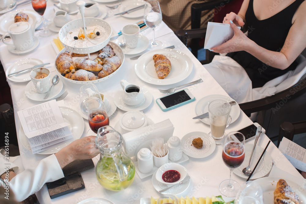 Crop view of people in elegant clothes sitting at long white restaurant table with beverages and pastries on dessert stands and plates from above