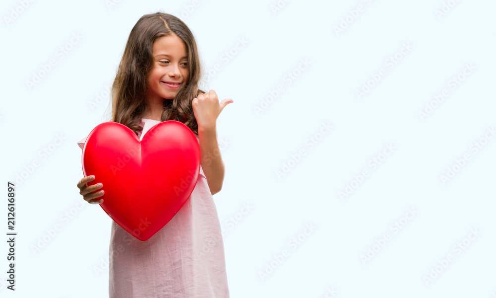 Brunette hispanic girl holding red heart pointing and showing with thumb up to the side with happy face smiling Brunette hispanic girl holding red heart pointing and showing with thumb up to the side with happy face smiling