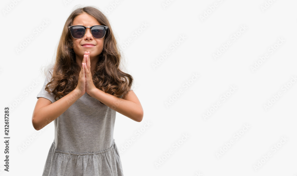Brunette hispanic girl wearing sunglasses begging and praying with hands together with hope expression on face very emotional and worried. Asking for forgiveness. Religion concept. Brunette hispanic girl wearing sunglasses begging and praying with hands together with hope expression on face very emotional and worried. Asking for forgiveness. Religion concept.