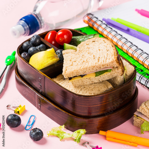 Healthy wooden lunch box with sandwiches, vegetables and fruits on pink background and school stationery. Childrens eating concept.