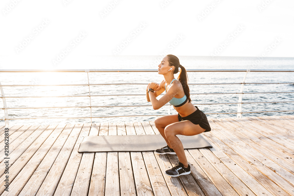 Photo of athletic caucasian woman 20s in tracksuit squatting, during workout on boardwalk at seaside - obrazy, fototapety, plakaty 