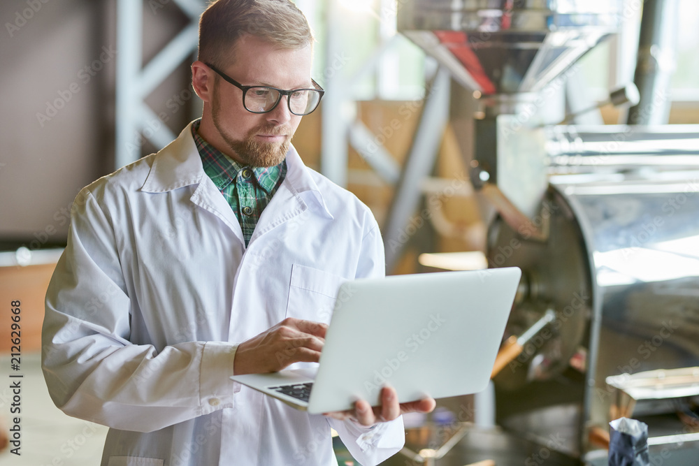 Waist up portrait of modern young man wearing lab coat using laptop while working in coffee production manufactory, copy space Waist up portrait of modern young man wearing lab coat using laptop while working in coffee production manufactory, copy space