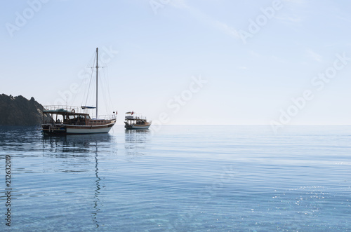 Small yacht and boat in the blue calm sea under cloudless sky in sunny morning; Sailboat with folded sails; Reflection of ships in calm water; Seascape with two boats