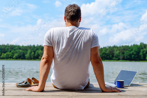 man sitting with laptop and looking on lake. working at vacation. summer time concept