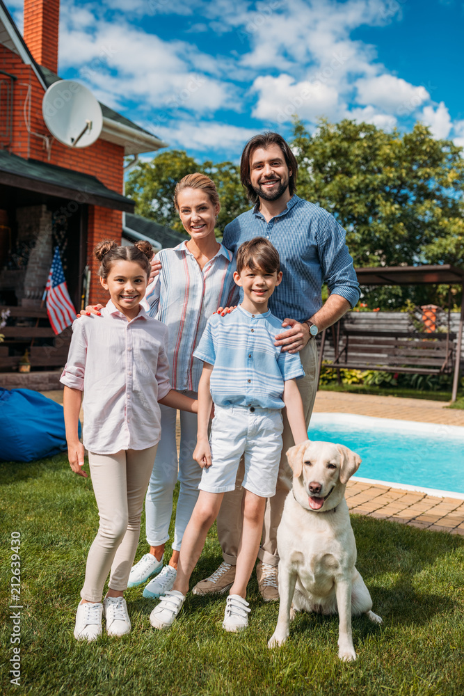 cheerful family with labrador dog looking at camera while standing on backyard of country house on summer day cheerful family with labrador dog looking at camera while standing on backyard of country house on summer day