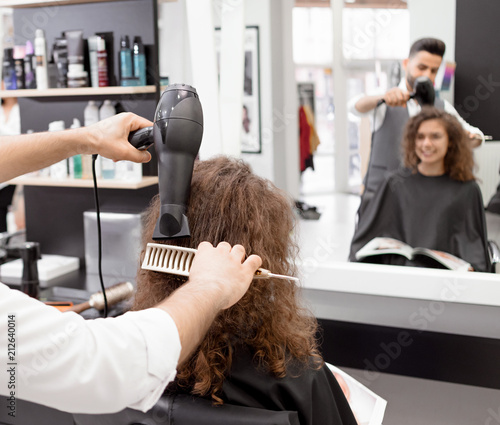 Backview of hairstyler drying female client's curly hair. Blurred reflection in big mirror. Using plastic brush and hair dryer. Wearing white shirt, grey waistcoat. modern stylish beatysalon.