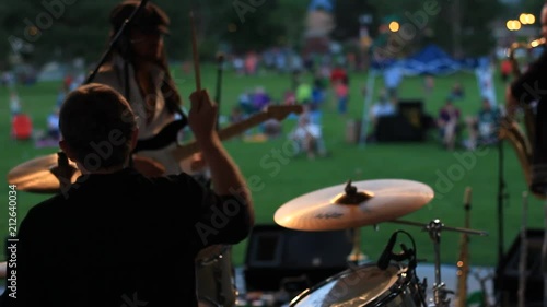 Out door concert, on stage a drummer and other musicians are performing for a crowd on the grass. photo