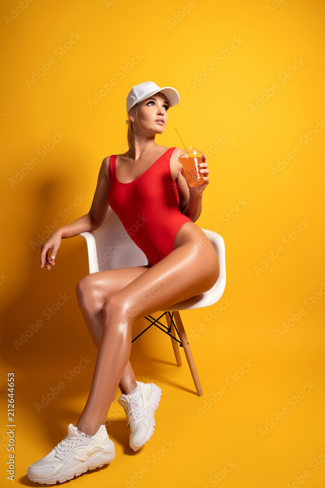 Luxury sexy woman in Red swimsuit, white baseball cap, white sneakers  sitting in chair in studio holding bottle and drinking beverage through  straw on yellow background foto de Stock | Adobe Stock