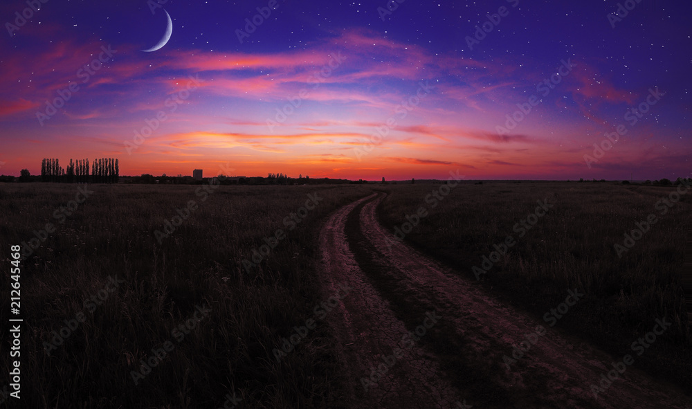 Sunset over the field with road at the village and young moon