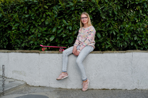 portrait of blond girl with pink cap and skateboard in park.  teenager girl in eyeglasses sitting outdoors on street.  Childhood, summer, vacation, sports  © skvalval