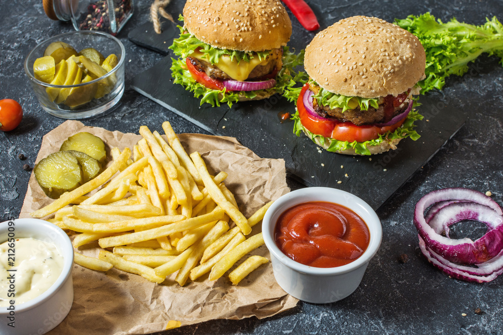 Two beef burgers, fries potatoes and cucumber on dark stone table Two beef burgers, fries potatoes and cucumber on dark stone table