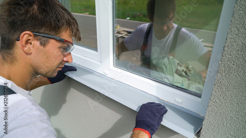 Man worker in safety glasses and protective gloves installing metal sill on external PVC window frame. Window sill installation process. Technology, exterior design, building concept photo