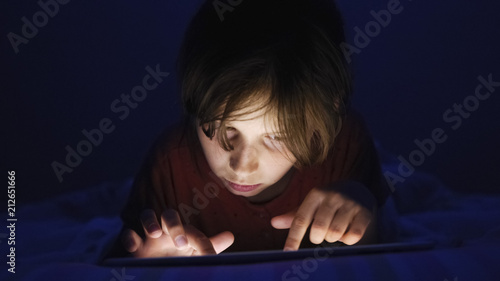 Dolly shot caucasian girl lying in bed playing tablet in social internet in dark light under blacket. Childhood, communication and technology concept