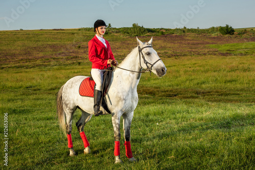 Young woman rider, wearing red redingote and white breeches, with her horse in evening sunset light. © nazarovsergey